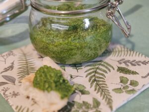 Rukkolás pesztó üvegben, mellette egy darabka kenyérre kenve, levélmintás szalvétán tálalva