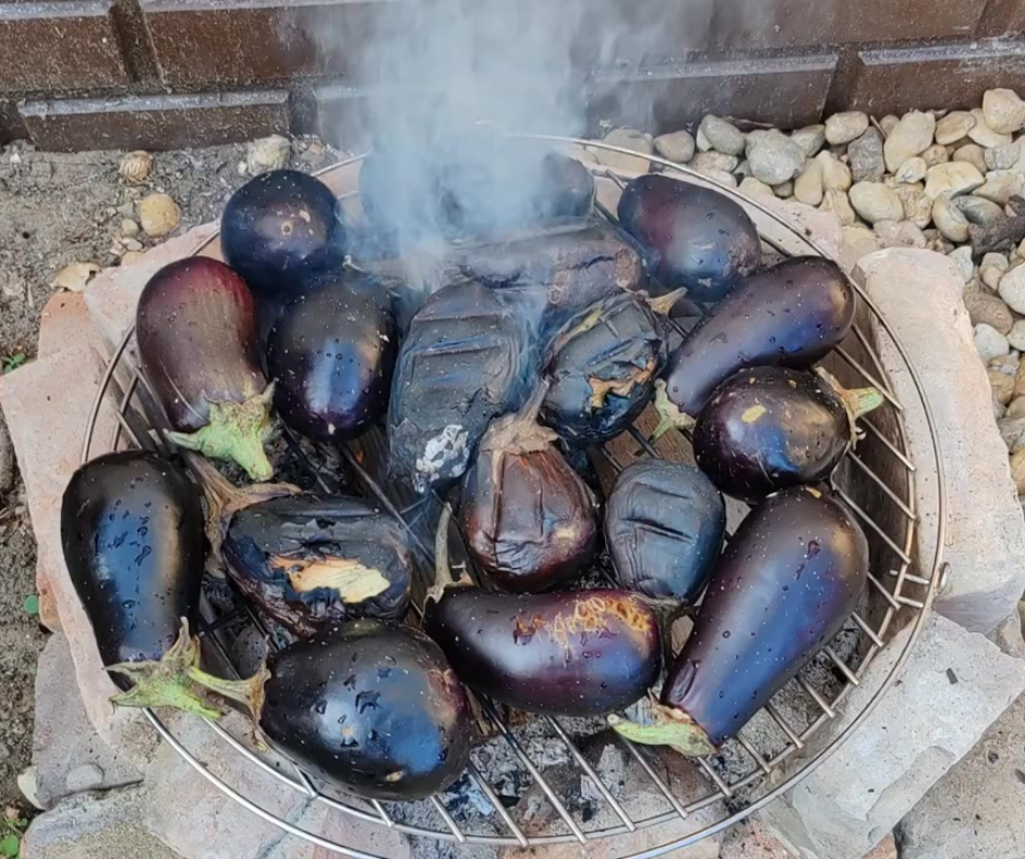 Padlizsánok grillezőrácson, szabad tűzön sülnek, a héjuk feketére ég, közben füst száll fel.