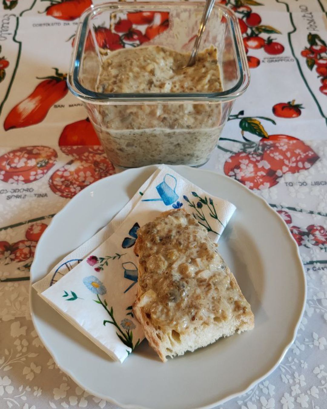 Házi padlizsánkrém üvegtálban, mellette egy szelet kenyérre kenve, virágmintás szalvétán, fehér tányéron.