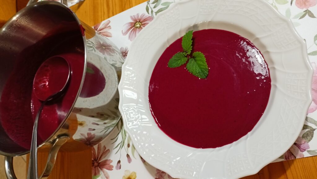 Fehér tányérban tálalt szederkrémleves, friss mentalevéllel díszítve, mellette egy fazékban merőkanál a levesből.