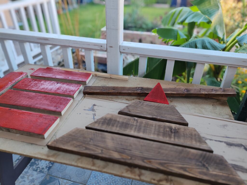 Festésre előkészített deszkák és a deszkákból összeállított karácsonyfa elemei egy asztalon, pirosra festett ládaoldalakkal és lazúrozott faágakkal, kertre néző háttérrel