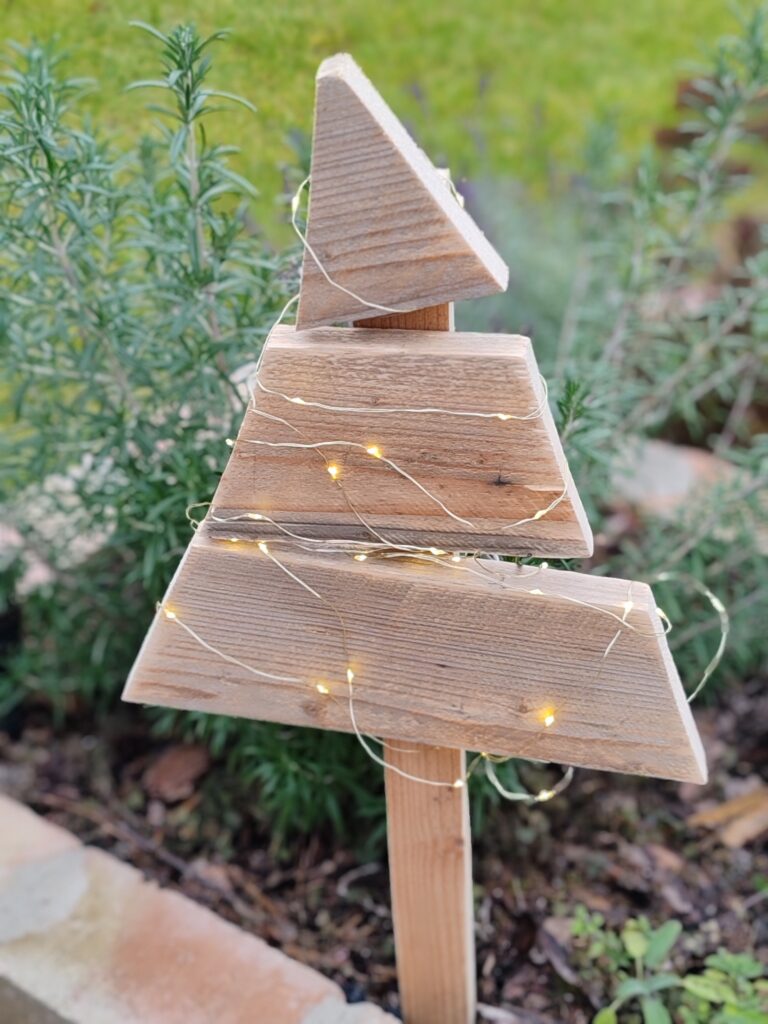 Natúr fából készült kültéri karácsonyfa mini égősorral díszítve, kerti növények előtt elhelyezve.