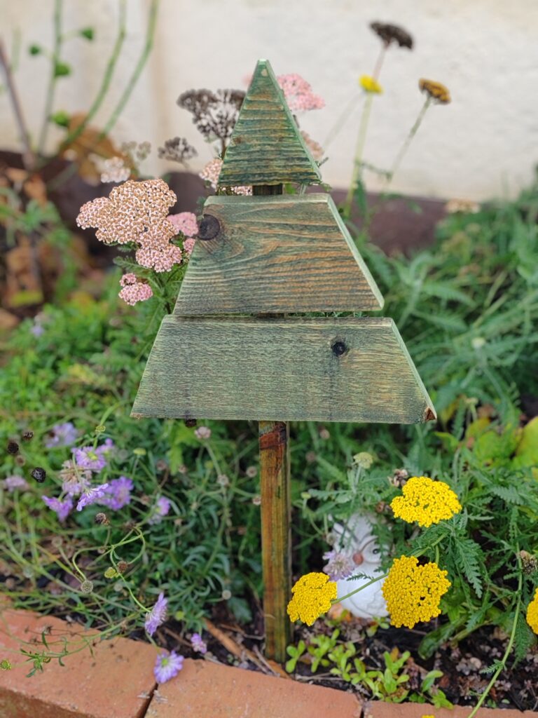 Zöldre festett, fából készült mini kültéri karácsonyfa virágágyásban, körülötte sárga, lila és rózsaszín virágokkal.