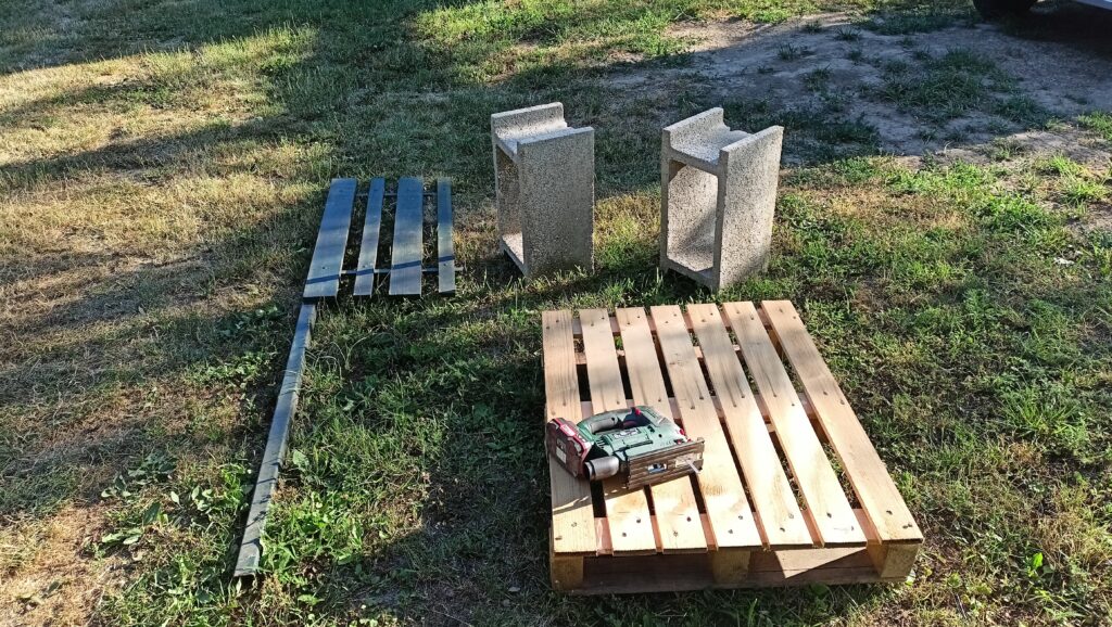 Raklap, két beton elem és több festett vaslemez a fűben, mellette egy vezeték nélküli fúró.