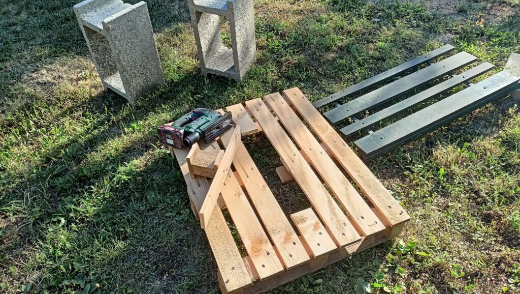 Raklap, amelyből egy rész ki van vágva, rajta deszkadarabok és egy vezeték nélküli fúró a fűben.