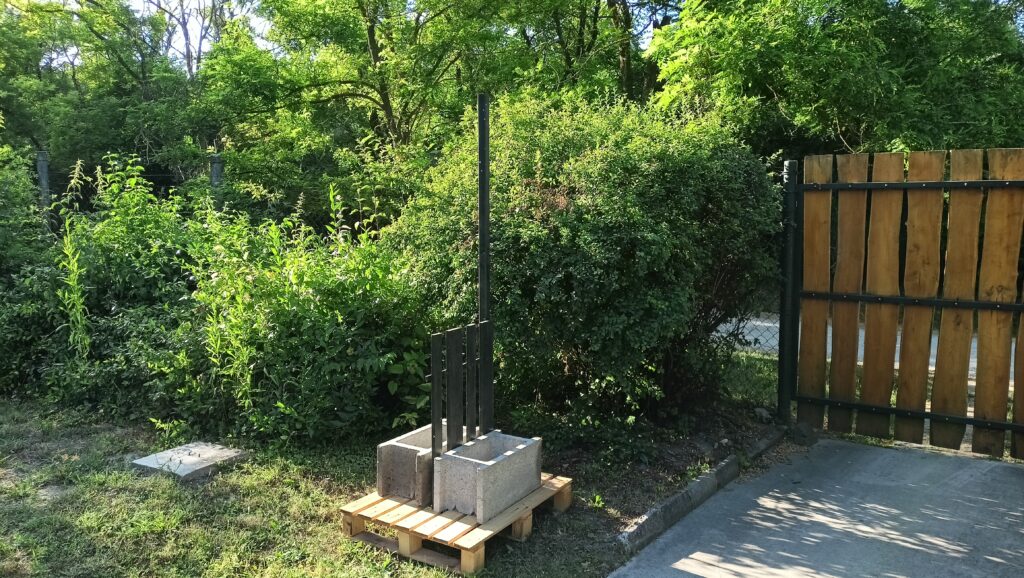 Raklapra helyezett két beton elem között áll egy magas, festett oszlop és mögötte egy kerítéselem, zöld bokrok és kapu mellett.