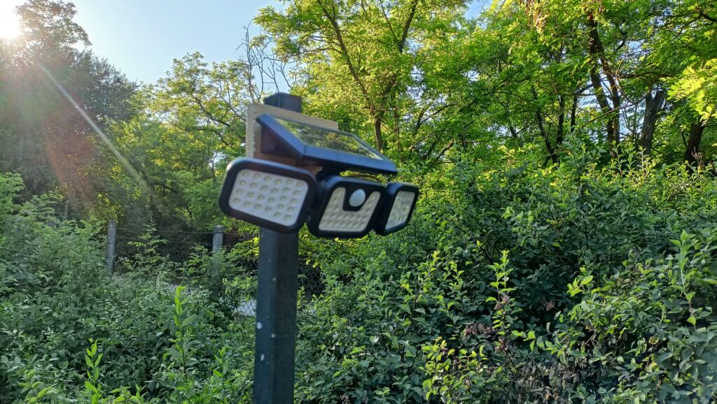 Kerti lámpaoszlop tetején napelemes, három paneles LED lámpa, háttérben zöld bokrokkal és fák között.