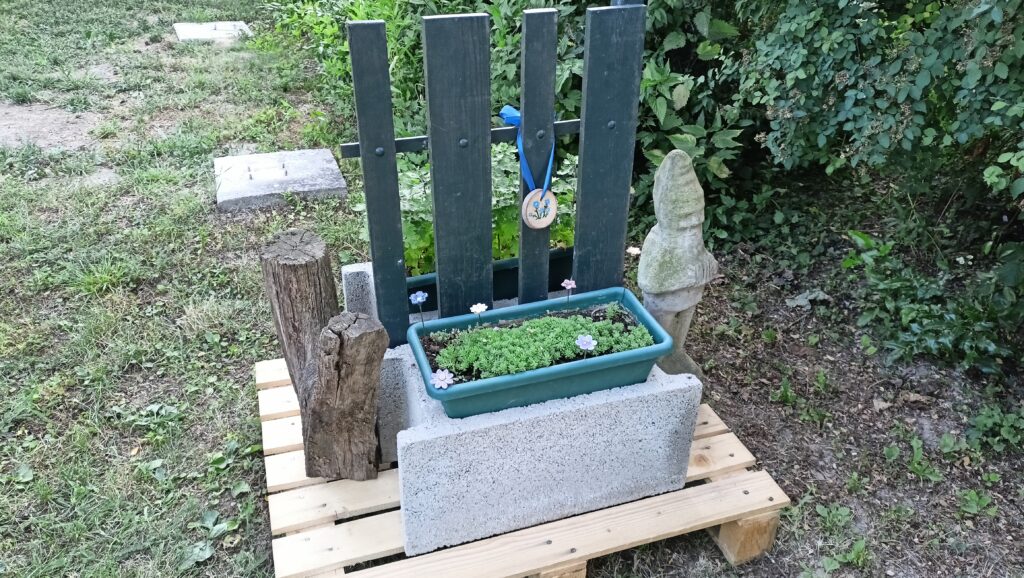 Növényláda pozsgásokkal, kis kerítéselem, fa tuskó és kerti figura a lámpaoszlop tövében, raklapra helyezve.