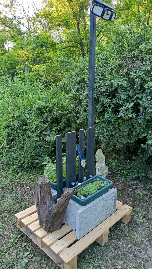 Kerti lámpaoszlop alap dekorral: raklap, beton elem, kis kerítéselem, növényláda és egy fa tuskó a kapu mellett.