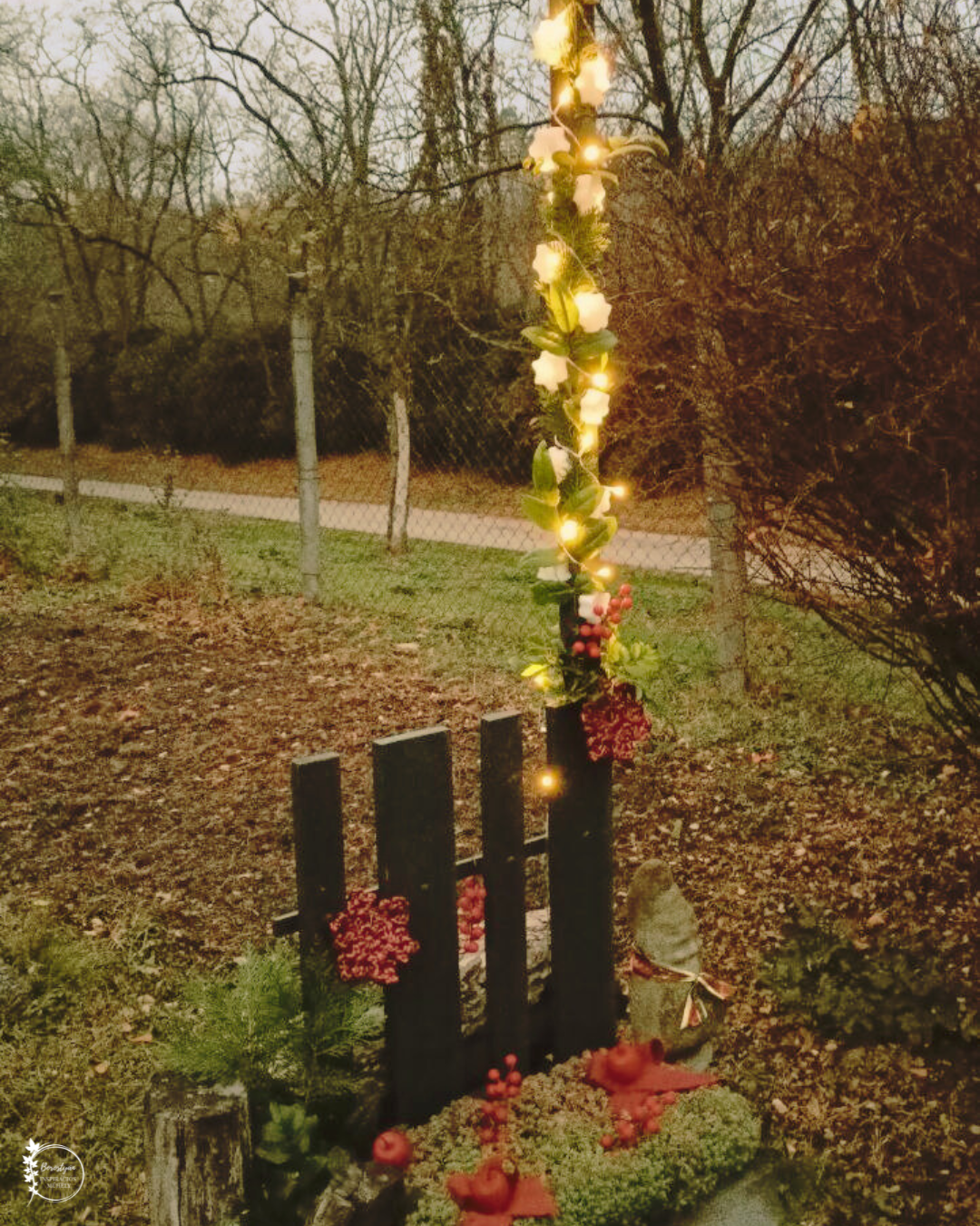 Kerti lámpaoszlop karácsonyi díszben: fényfüzér, örökzöld és piros dekor a kapu mellett, esti fényekben.