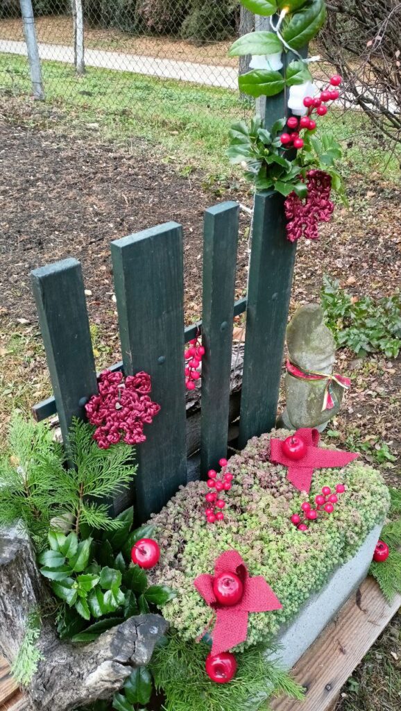 Karácsonyi dekor a lámpaoszlop tövében: örökzöld ágak, piros almadíszek, bogyók és masnik egy beton elem és kerítés előtt.