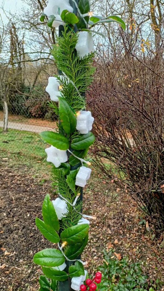 Lámpaoszlopra tekert fényfüzér, örökzöld ágak és fehér dekor elemek, téli kerti háttérrel.