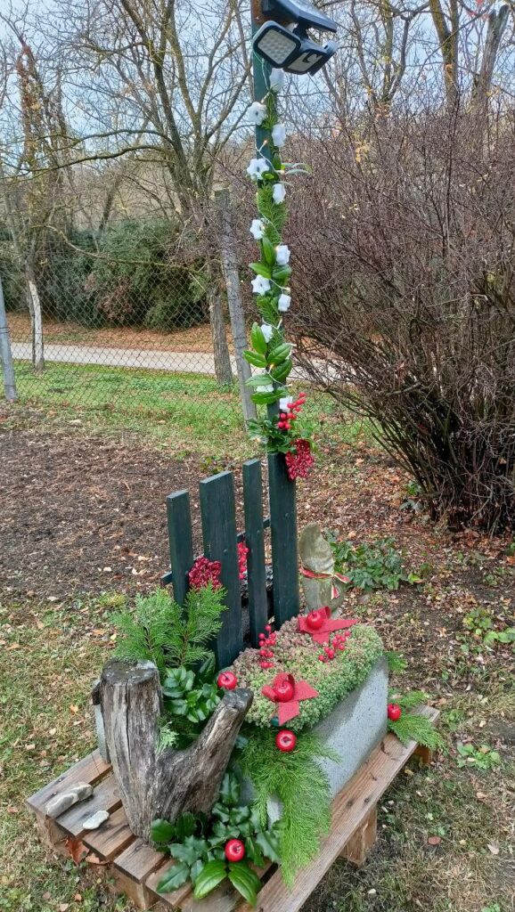 Karácsonyi díszbe öltöztetett kerti lámpaoszlop beton alappal, örökzölddel, piros almákkal és masnikkal a kapu mellett.